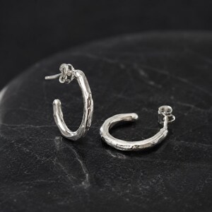 May include: A pair of silver hoop earrings. The earrings have a textured, organic design. The posts are visible, and the earrings are set against a dark background. The earrings are a simple, elegant accessory.