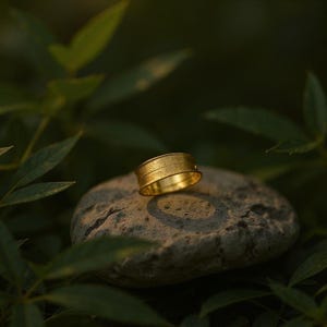 May include: A gold ring with a textured surface, resting on a gray stone. The ring has a band design with a subtle, linear pattern. The background features blurred green foliage, creating a natural and elegant setting for the jewelry.