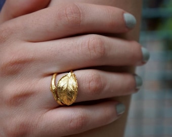 Puede incluir: Un anillo dorado con diseño de hoja. El anillo presenta una forma de hoja texturizada y detallada como pieza central, con una banda delicada. El anillo se lleva en un dedo, mostrando su intrincado diseño y brillo metálico.
