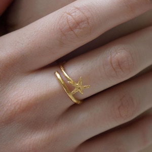 Peut inclure: Une bague dorée avec un motif d'étoile de mer. La bague a un design à double bande avec un petit charme d'étoile de mer. La bague est portée sur un doigt, avec la main dans l'image.