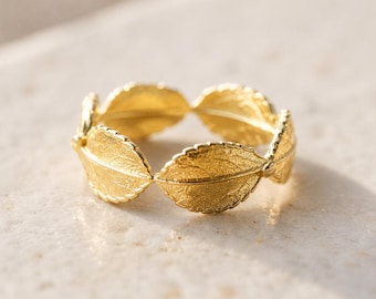 Puede incluir: Un anillo dorado con un diseño de hojas repetidas. Las hojas tienen vetas detalladas y una superficie texturizada. El anillo se muestra sobre un fondo neutro, resaltando su intrincado diseño.