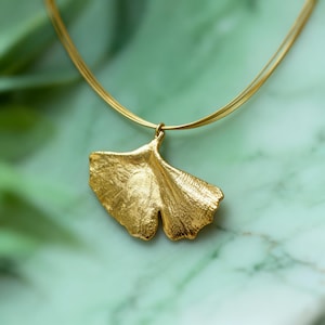 Può includere: Una collana color oro con un ciondolo a forma di foglia di ginkgo. Il ciondolo presenta una superficie metallica testurizzata con una caratteristica silhouette a ventaglio. È sospeso a una sottile e delicata catena in filo color oro.