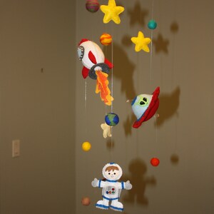 May include: A colourful felt mobile with a rocket, a UFO, an astronaut, and stars hanging from a blue ring. The mobile is made of felt and has a variety of colours, including red, blue, green, yellow, and orange.