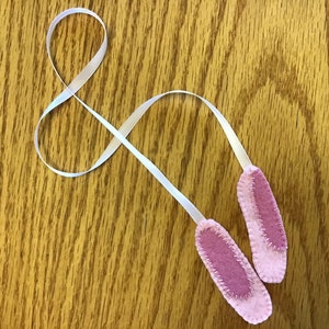 Pode incluir: Duas orelhas de coelho de feltro rosa com fita branca, sobre uma superfície de madeira. As orelhas têm formato oval com interior rosa claro e costuras brancas. A fita é fina e branca.