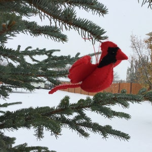 Peut inclure: Ornement fait main représentant un cardinal rouge avec des détails noirs, suspendu à une branche de pin. L'oiseau est accroché par une ficelle rouge sur un fond enneigé, évoquant l'hiver ou les fêtes.