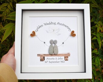 May include: A white framed picture with a golden wedding anniversary message. The picture features a couple made of stones, a dove, and two golden hearts. The text reads "Golden Wedding Anniversary Annette & John 16th September 1972".