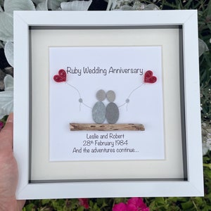 Peut inclure: Une image encadrée blanche avec un design d'art de galets représentant deux personnages se tenant la main. Les personnages sont assis sur un petit morceau de bois flotté. Le texte "Ruby Wedding Anniversary" est en haut de l'image. Le texte "Leslie and Robert 28th February 1984 And the adventures continue..." est en bas de l'image.