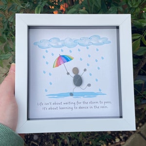 May include: Framed pebble art depicting a figure dancing in the rain with a rainbow umbrella. The artwork includes blue clouds and raindrops. The text reads, "Life isn't about waiting for the storm to pass, It's about learning to dance in the rain." The frame is white.