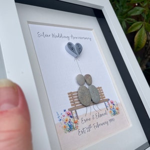 May include: A framed artwork featuring a silver wedding anniversary celebration. Two pebble figures sit on a bench with a paper heart balloon above them. The artwork includes the text "Silver Wedding Anniversary" and "Esme & Edward Est 28th February 1994".