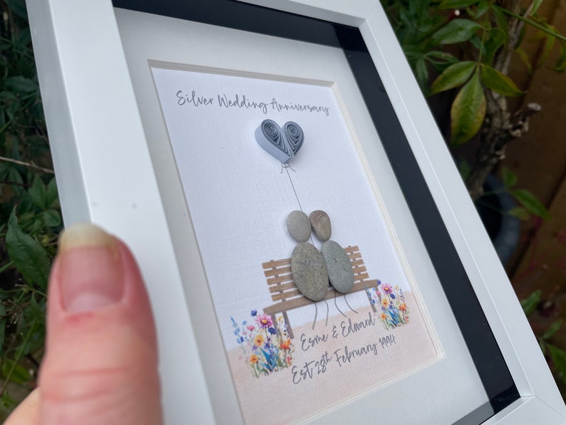 May include: A framed artwork featuring a silver wedding anniversary celebration. Two pebble figures sit on a bench with a paper heart balloon above them. The artwork includes the text "Silver Wedding Anniversary" and "Esme & Edward Est 28th February 1994".