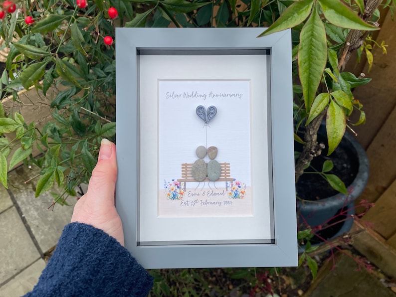 May include: A framed pebble art picture of a couple sitting on a bench with a heart-shaped balloon. The text reads "Silver Wedding Anniversary" and "Eve & Edmond Est 28th February 1993".