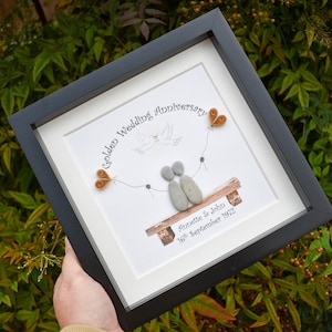 May include: A framed pebble art piece celebrating a golden wedding anniversary. The art features two figures made of pebbles sitting on a bench with a dove flying above them. The text reads "Golden Wedding Anniversary" and "Annette & John 16th September 1972".