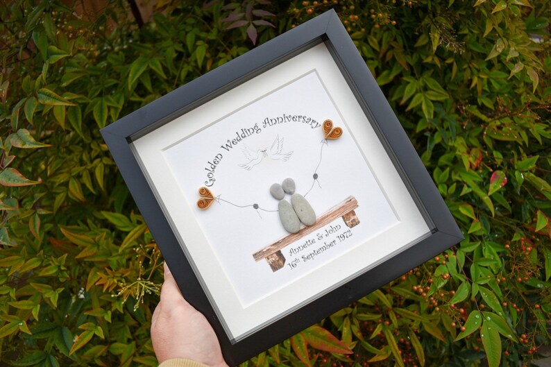 May include: A framed pebble art piece celebrating a golden wedding anniversary. The art features two figures made of pebbles sitting on a bench with a dove flying above them. The text reads "Golden Wedding Anniversary" and "Annette & John 16th September 1972".