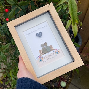May include: A framed picture with a silver wedding anniversary theme. The picture shows two stone figures sitting on a bench with a heart-shaped balloon above them. The text reads "Silver Wedding Anniversary" and "Esme & Edward Est 2nd February 1994".