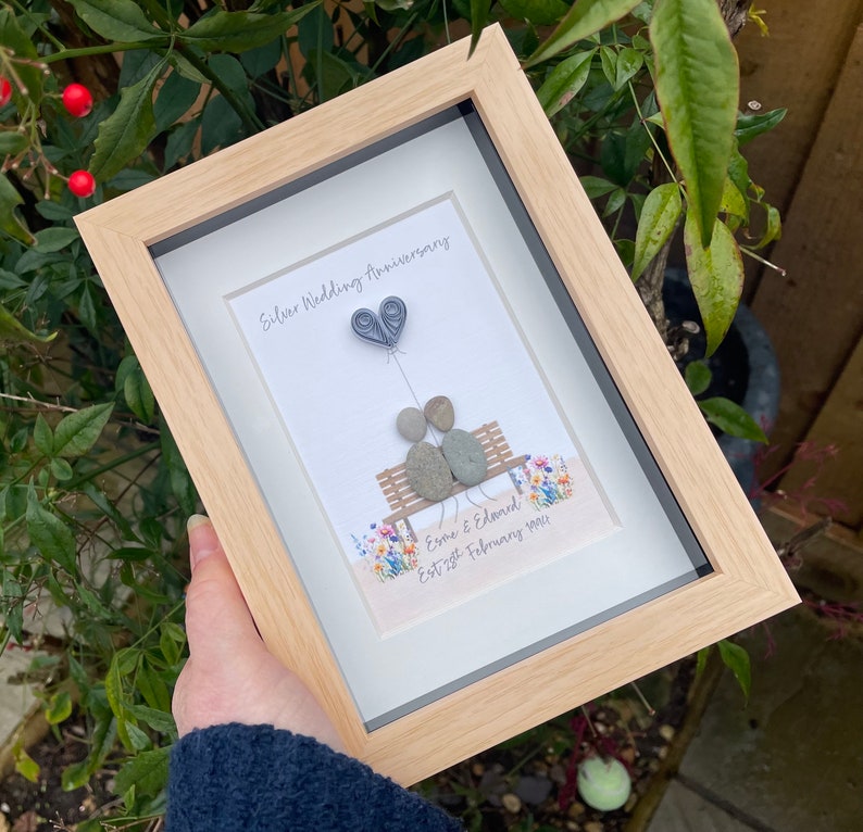 May include: A framed picture with a silver wedding anniversary theme. The picture shows two stone figures sitting on a bench with a heart-shaped balloon above them. The text reads "Silver Wedding Anniversary" and "Esme & Edward Est 2nd February 1994".