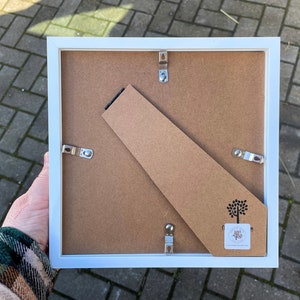 May include: White picture frame with a cardboard backing and a small tree logo. The frame has metal hanging hardware on the back.