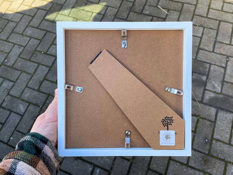 May include: White picture frame with a cardboard backing and a small tree logo. The frame has metal hanging hardware on the back.
