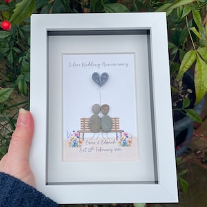 May include: A white framed picture with a silver wedding anniversary message. The picture features a couple made of stones sitting on a bench with a heart balloon above them. The text reads "Silver Wedding Anniversary" and "Esme & Edward Est 25th February 1998".