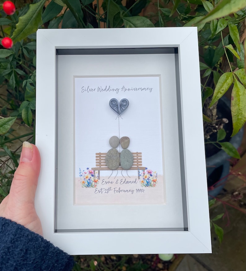 May include: A white framed picture with a silver wedding anniversary message. The picture features a couple made of stones sitting on a bench with a heart balloon above them. The text reads "Silver Wedding Anniversary" and "Esme & Edward Est 25th February 1998".