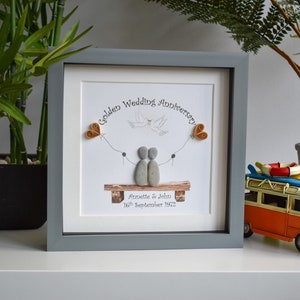 May include: A framed pebble art picture with a gray frame. The picture shows two figures made of pebbles sitting on a bench with a dove flying above them. The text on the picture reads "Golden Wedding Anniversary Annette & John 16th September 1972".