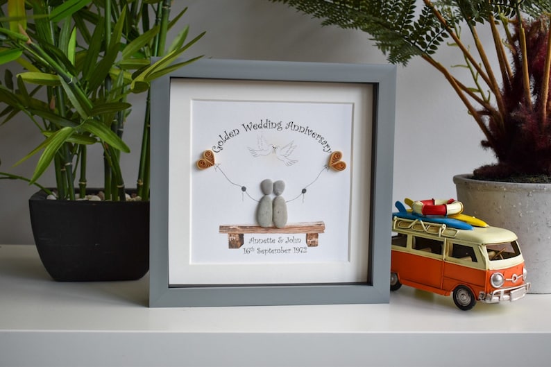 May include: A framed pebble art picture with a gray frame. The picture shows two figures made of pebbles sitting on a bench with a dove flying above them. The text on the picture reads "Golden Wedding Anniversary Annette & John 16th September 1972".