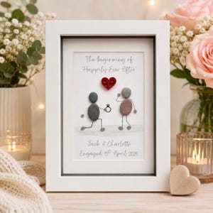 May include: A white framed wall art piece featuring pebble art of two figures, one kneeling and offering a ring to the other. Above them is a red paper quilled heart and the text 'The beginning of 'Happily Ever After'', 'Jack & Charlotte', and 'Engaged 1st April 2025'.