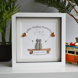 May include: A white framed picture with a golden wedding anniversary message. The picture features a couple made of stones, a dove, and two golden hearts. The text reads "Golden Wedding Anniversary Annette & John 16th September 1972".