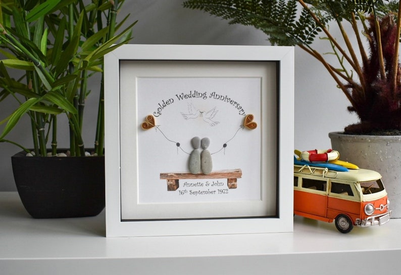 May include: A white framed picture with a golden wedding anniversary message. The picture features a couple made of stones, a dove, and two golden hearts. The text reads "Golden Wedding Anniversary Annette & John 16th September 1972".