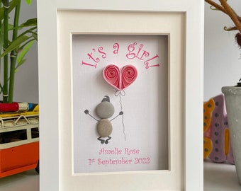 May include: A white picture frame with a pink and white pebble art design. The art features a stick figure holding a pink heart balloon. The text "It's a girl!" is written in pink letters. The text "Amelie Rose 1st September 2022" is written in pink letters below the art.