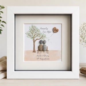 May include: A square white frame containing a pebble art piece featuring two figures made of smooth gray and brown stones sitting on a bench under a painted tree. The text reads Happily Ever After, Aimee & Barry, and 8th August 2026.