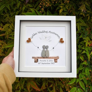 May include: A white framed picture with a golden wedding anniversary message. The picture features a couple made of stones sitting on a bench with a dove flying above them. The couple is holding hands and there are two small orange hearts on strings above them. The text reads "Golden Wedding Anniversary" and "Annette & John 16th September 1972".