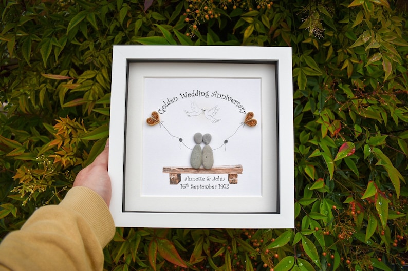 May include: A white framed picture with a golden wedding anniversary message. The picture features a couple made of stones sitting on a bench with a dove flying above them. The couple is holding hands and there are two small orange hearts on strings above them. The text reads "Golden Wedding Anniversary" and "Annette & John 16th September 1972".