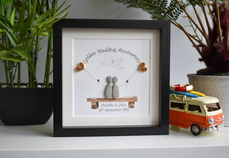 May include: A framed pebble art piece with a couple sitting on a bench. The art is titled "Golden Wedding Anniversary" and includes the names Annette & John and the date 16th September 1972.