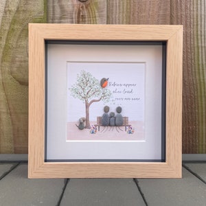 Op de afbeelding: Ingelijste kunst met kiezelstenen die een gezin en een hond op een bank onder een boom afbeeldt. Een roodborstje zit op de boom. De tekst luidt: "Robins appear when loved ones are near."