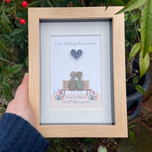 May include: A framed pebble art picture with a silver wedding anniversary theme. The picture shows two pebble people sitting on a bench with a heart balloon above them. The text reads "Silver Wedding Anniversary" and "Esme & Edward Est 28th February 1995".