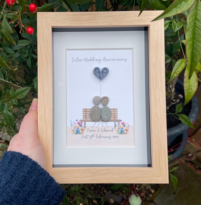 May include: A framed pebble art picture with a silver wedding anniversary theme. The picture shows two pebble people sitting on a bench with a heart balloon above them. The text reads "Silver Wedding Anniversary" and "Esme & Edward Est 28th February 1995".