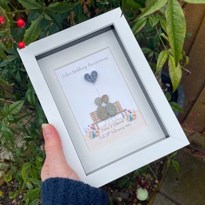 May include: A white frame with a white mat and a picture of two gray stones representing a couple sitting on a bench with a gray paper heart balloon. The text "Silver Wedding Anniversary" is at the top of the picture and the text "Esme & Edward Est 27th February 1994" is at the bottom of the picture.