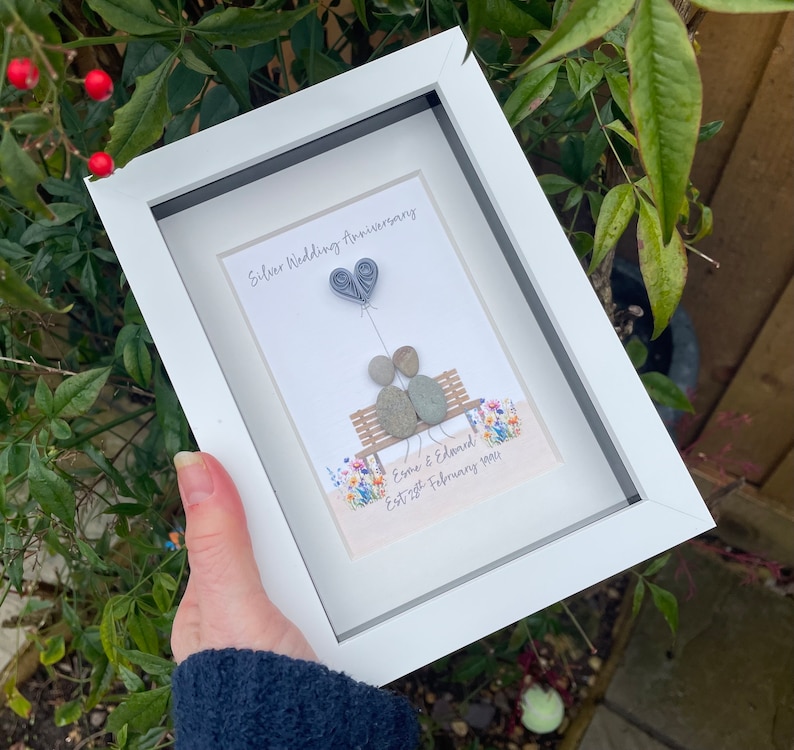 May include: A white frame with a white mat and a picture of two gray stones representing a couple sitting on a bench with a gray paper heart balloon. The text "Silver Wedding Anniversary" is at the top of the picture and the text "Esme & Edward Est 27th February 1994" is at the bottom of the picture.
