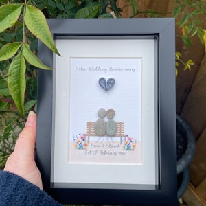 May include: A framed pebble art piece depicting a couple sitting on a bench with a heart-shaped balloon. The text reads "Silver Wedding Anniversary, Esme & Edward Est. 28th February 1994".