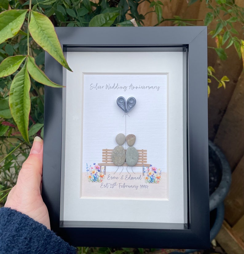 May include: A framed pebble art piece depicting a couple sitting on a bench with a heart-shaped balloon. The text reads "Silver Wedding Anniversary, Esme & Edward Est. 28th February 1994".