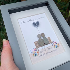 May include: A framed artwork featuring a couple made of stones sitting on a bench with a paper heart balloon above them. The artwork is decorated with a floral pattern and the text "Silver Wedding Anniversary, Esme & Edward Est 28th February 1994".