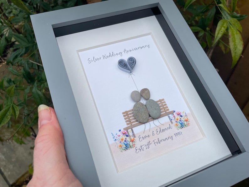 May include: A framed artwork featuring a couple made of stones sitting on a bench with a paper heart balloon above them. The artwork is decorated with a floral pattern and the text "Silver Wedding Anniversary, Esme & Edward Est 28th February 1994".