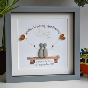 May include: A framed pebble art piece celebrating a golden wedding anniversary. The art features two pebble figures holding hands, sitting on a bench with a dove flying above them. The text reads "Golden Wedding Anniversary" and "Annette & John 16th September 1972".