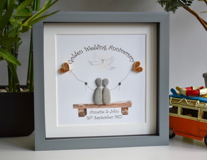 May include: A framed pebble art piece celebrating a golden wedding anniversary. The art features two pebble figures holding hands, sitting on a bench with a dove flying above them. The text reads "Golden Wedding Anniversary" and "Annette & John 16th September 1972".