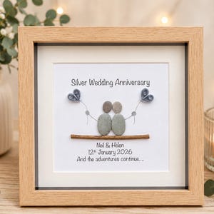 May include: A square framed wall art piece featuring pebble art of two figures sitting on a twig. The text reads Silver Wedding Anniversary, Neil & Helen, 12th January 2026, And the adventures continue. Two blue paper quilled hearts are attached.
