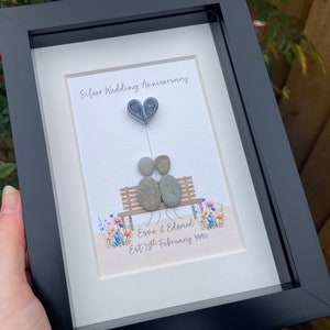 May include: A framed pebble art piece depicting a couple sitting on a bench with a paper heart balloon. The text reads "Silver Wedding Anniversary, Esme & Edward, Est 28th February 1994".