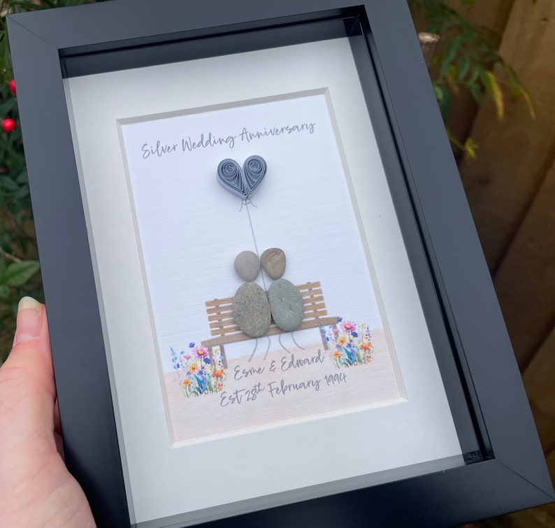 May include: A framed pebble art piece depicting a couple sitting on a bench with a paper heart balloon. The text reads "Silver Wedding Anniversary, Esme & Edward, Est 28th February 1994".