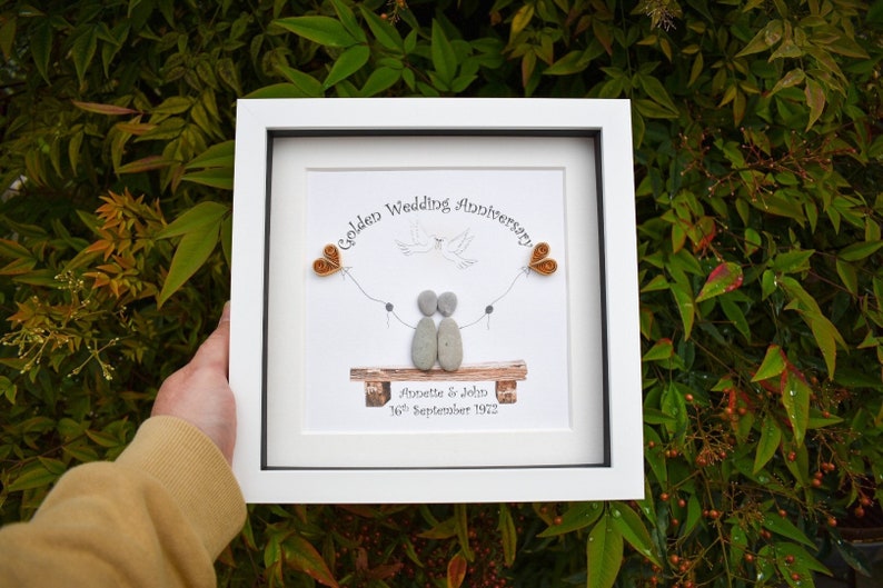 May include: A white framed picture with a golden wedding anniversary message. The picture features a couple made of stones, a dove, and two golden hearts. The text reads "Golden Wedding Anniversary Annette & John 16th September 1972".