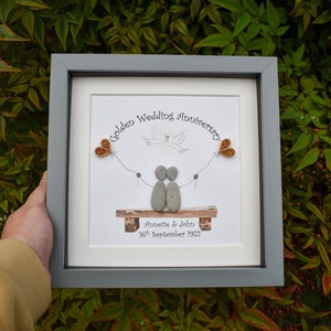 May include: A framed pebble art piece depicting a couple sitting on a bench with two hearts and a dove flying above them. The text reads "Golden Wedding Anniversary Annette & John 16th September 1972".