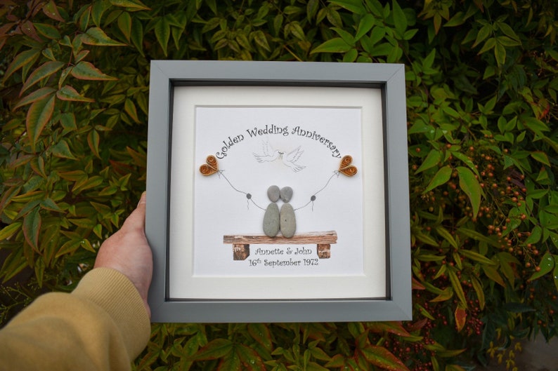 May include: A framed pebble art piece depicting a couple sitting on a bench with two hearts and a dove flying above them. The text reads "Golden Wedding Anniversary Annette & John 16th September 1972".